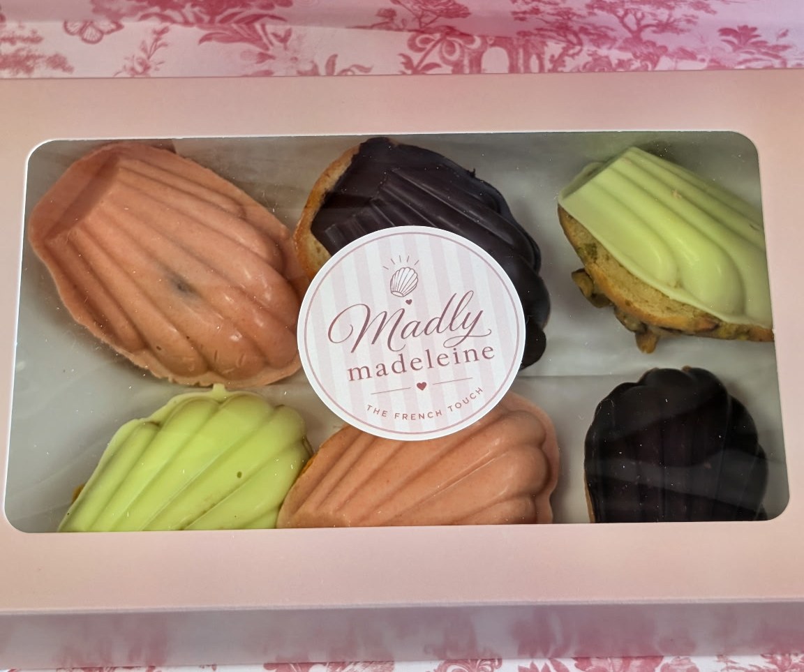 A pink Madly Madeleine gift box of six madeleines with the logo sticker, beside a small sauce cup