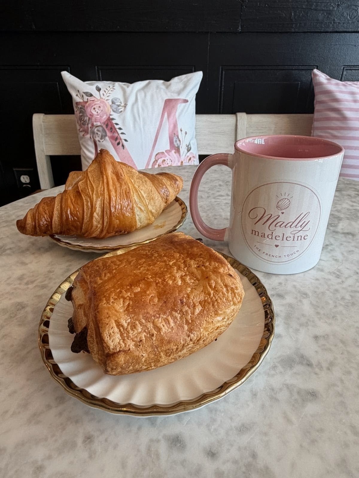 A croissant and a chocolatine on plates beside a Madly Madeleine mug on a marble table in the café